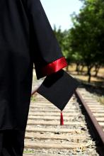 Graduate in black gown holding black mortarboard to side walking along train tracks