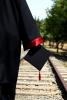 Graduate in black gown holding black mortarboard to side walking along train tracks