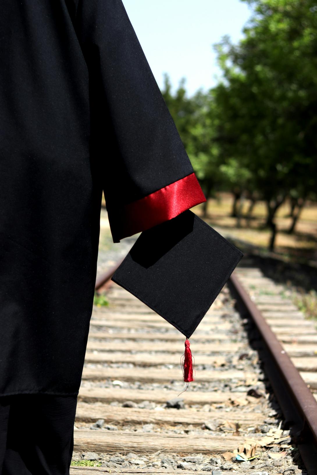 Graduate in black gown holding black mortarboard to side walking along train tracks